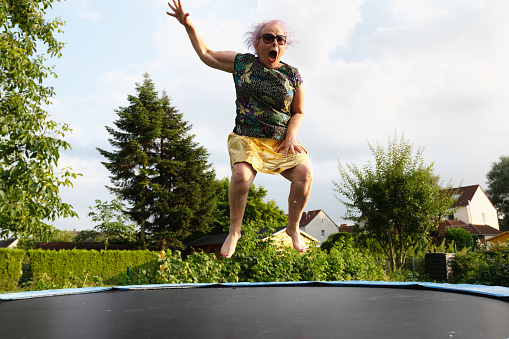Eccentric senior woman with dyed pink hair, wearing a black shirt with colorful pattern and a golden skirt, jumping on trampoline