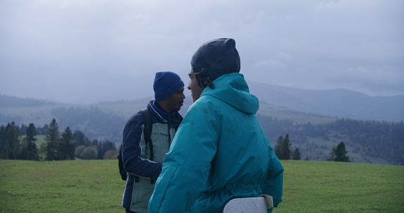 Two African American tourists stand on top of hill and admire scenery. Group of travelers stopped to rest after long walking trek on mountains. Concept of nature discovery and tourism. Active leisure.