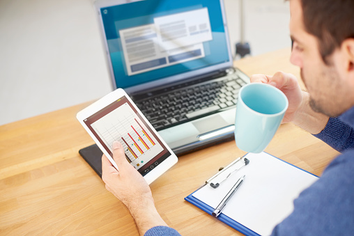 Rear view shot of young businessman holding digital tablet in his hand and analyzing financial data while sitting at desk in front of laptop and drinking coffee. Focus on foreground.