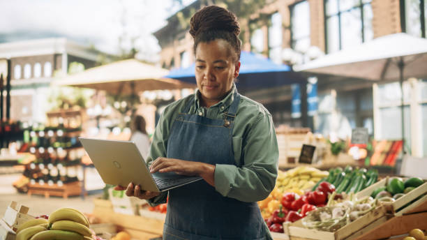 istock African Farmers Market Manager Using Laptop Computer while Standing Next to Street Vendor Stalls with Fresh Organic Agricultural Products. Middle Aged Black Female Managing Business Operations 1934640812