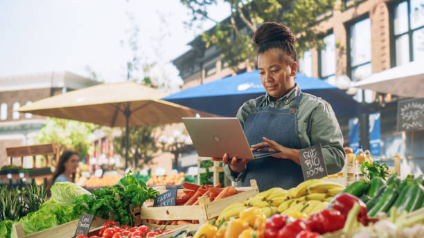 successful female farmer working on a laptop computer, catching up with supplier online to plan future orders for her organic street vendor stall with sustainable fruits and vegetables - farmer laptop selling internet foto e immagini stock