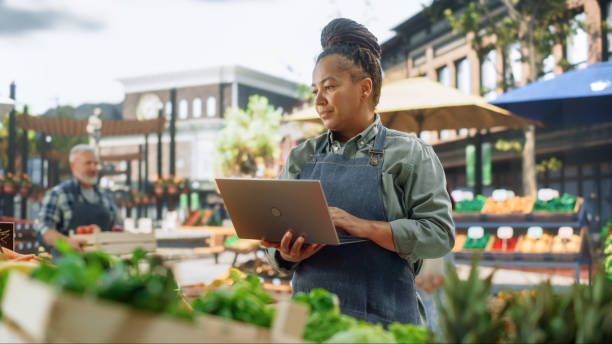 istock African Farmer Using Laptop Computer on a City Square with Vendors Selling Fresh Organic Agricultural Products. Middle Aged Black Female Managing Business Operations and Communications with Suppliers 1934653259