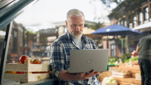 portrait of a middle aged street vendor working on laptop computer while sitting in a farmers market stall with fresh natural agricultural products. businessman contacting supply stores online - farmer laptop selling internet стоковые фото и изображения