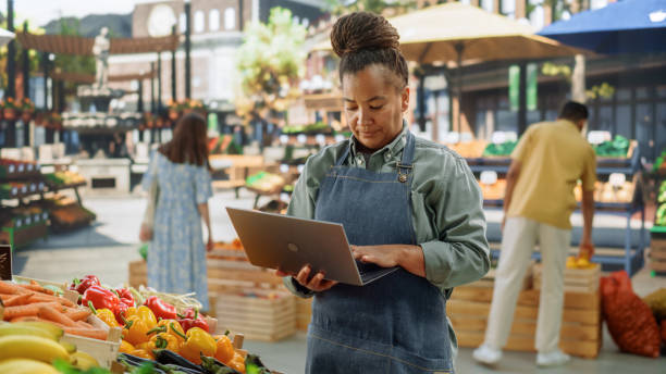istock African Farmer Using Laptop Computer on a City Square with Vendors Selling Fresh Organic Agricultural Products. Middle Aged Black Female Managing Business Operations and Communications with Suppliers 1934671574