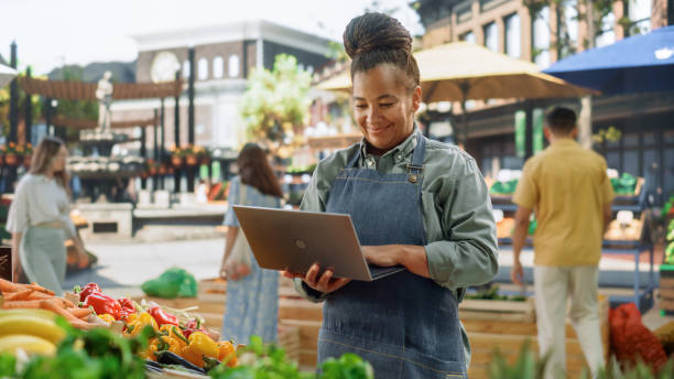 istock African Farmer Using Laptop Computer on a City Square with Vendors Selling Fresh Organic Agricultural Products. Middle Aged Black Female Managing Business Operations and Communications with Suppliers 1934671585