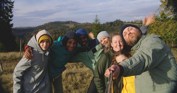 POV from phone front camera. Caucasian man, blogger with selfie stick records video with multiethnic boys on top of the hill and talks to camera. Group of diverse travelers during hike in mountains.