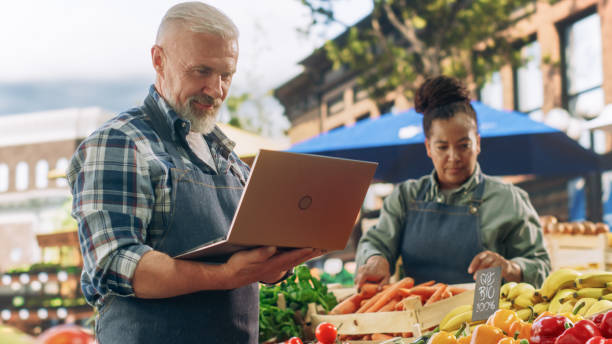istock Market Manager Using Laptop Computer while Standing Next to Street Vendor Stalls with Fresh Organic Agricultural Products. Adult Male Managing Business Development in Rural Farmers Community 1934737687