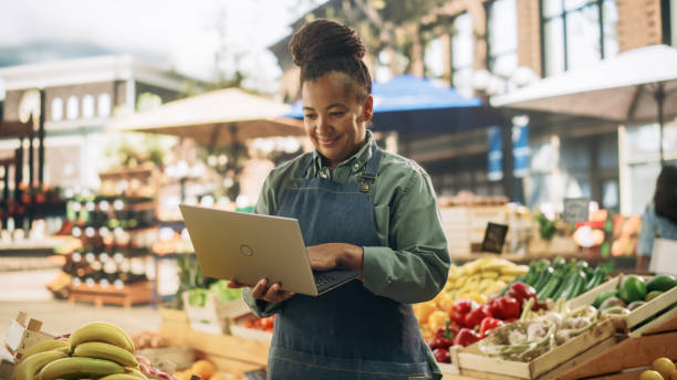 istock African Farmers Market Manager Using Laptop Computer while Standing Next to Street Vendor Stalls with Fresh Organic Agricultural Products. Middle Aged Black Female Managing Business Operations 1934741104