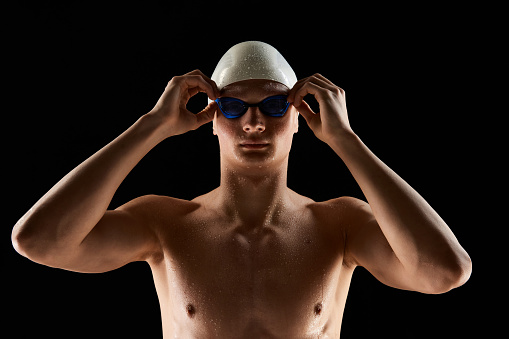 Portrait of young man, swimmer wearing goggles and swimming cap isolated over black background. Motivated aquatic athlete. Poster for sport schools, events. Concept of competition, tournament