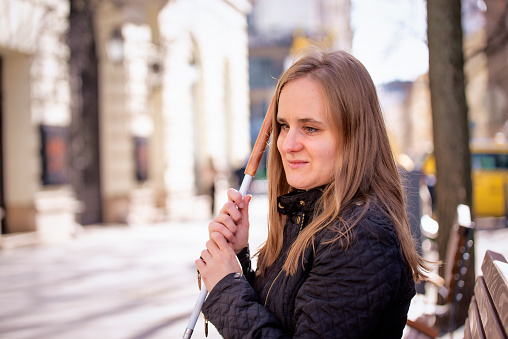 Close-up of a visually impaired woman holding a white cane and sitting on a bench in the city. Smiling female wearing casual clothes.