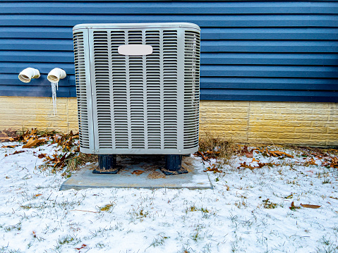 An electric heat pump and ac unit is outside a blue vinyl sided house in the winter time. There is snow on the ground. There are two exhaust pipes for the internal unit. Icicles hang from one of them.