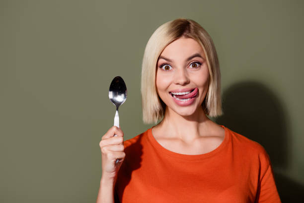 портрет девушки, высунувшей язык, лижет зубы, держа серебряную ложку, в ожидании лукового супа в ресторане, изолированном на фоне цвета хаки - silver spoon in mouth стоковые фото и изображения