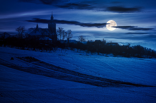church in the village in winter at night. rural landscape on the snow covered hill in full moon light
