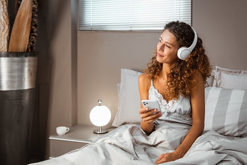 Young calm female wearing nightwear sitting in bed using smartphone while listening to music on headphones from playlist in bedroom