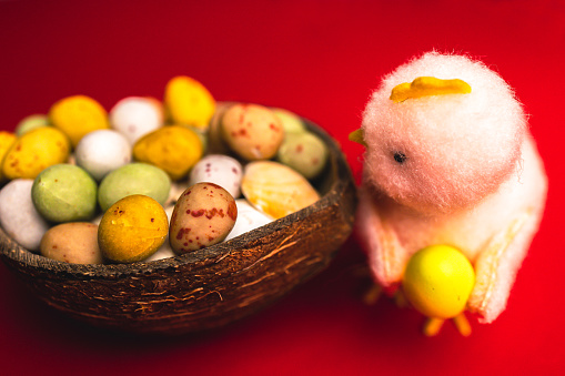 The image exudes the playful charm of Easter with a soft, plush chick standing guard over a coconut shell brimming with multicolored candy eggs. Set against a striking red background, the chick appears almost lifelike, with a little halo and wings, suggesting a whimsical Easter angel. The candy eggs, in hues of yellow, green, and cream, speckled with red, add a sweet touch to the festive scene.