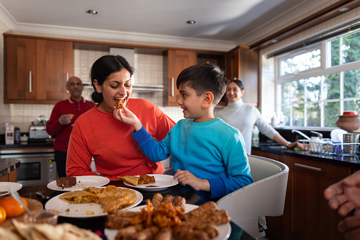 A waist-up shot of a multigenerational family in the kitchen. The young boy is feeding his mother a snack. The family are all wearing casual clothing located in Middlesborough, England.