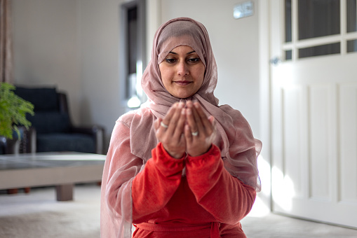 Waist-up shot of a mid-adult woman praying at home looking down at her hands sitting- kneeling in the living room in her family home. She is wearing traditional headwear. Located in Middlesborough, England.