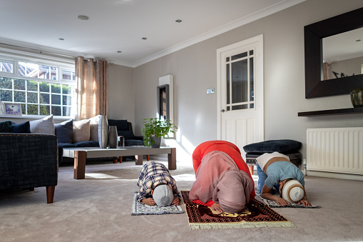 Full shot of a mother and her two children praying at home on prayer mats side by side in the living room of their family home. They are wearing traditional headpieces and casual clothing. Located in Middlesborough, England.