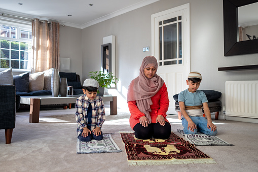 Full shot of a mother and her two children praying at home on prayer mats side by side in the living room of their family home. They are wearing traditional headpieces and casual clothing. Located in Middlesborough, England.