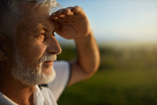 Bearded man squinting from bright sun, while looking into distance during trip. Senior bearded man using hand to protect eyes from sun, smiling outside. Close up view of male tourist squinting from bright light, while looking into distance, during mountain trip. Concept of watch. squint eyes stock pictures, royalty-free photos & images