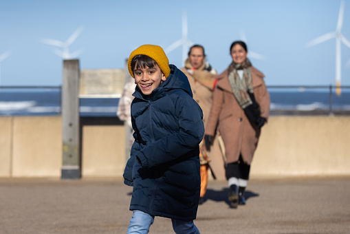A three-quarter length shot of a multigenerational family walking and laughing together wearing warm clothing. Their child is running ahead of the adults smiling to himself. They are located on a public footpath alongside Redcar Beach near Middlesborough.