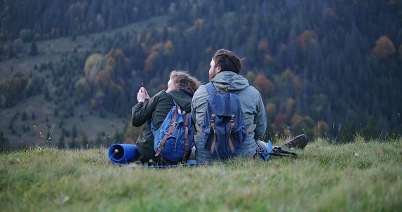 Happy Caucasian couple of hikers sits on the hill, looks at photos on the phone or surfs the Internet. Family of travelers during hiking trip or trek in the mountains. Tourism and outdoor recreation.