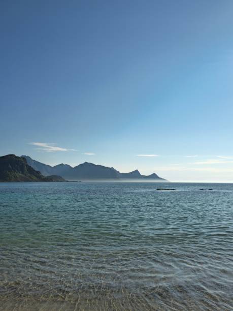Pristine white sand stretches out to meet turquoise waters as dramatic, snow-capped mountains rise in the distance. This is Haukland Beach, a jewel of the Lofoten Islands in Norway, where the Arctic landscape transforms into a tropical paradise. The sun bathes the scene in a warm glow, inviting visitors to swim, sunbathe, or hike the surrounding peaks.