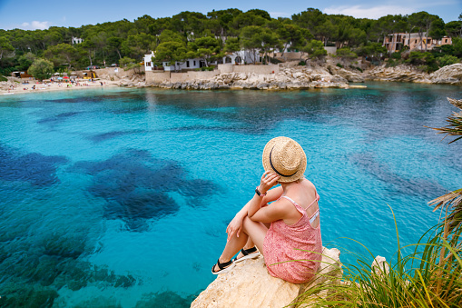 Rear view of a woman sitting on a cliff, admiring the coastal view of Cala Gat beach in Mallorca, Spain