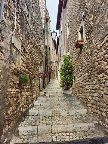 A narrow, winding alleyway paved with cobblestones in the quaint old town of Sermoneta, Italy. The alley curves out of sight, framed by tall stone buildings with red tile roofs and colorful window boxes overflowing with flowers. Sunlight streams through a gap between the buildings, illuminating the damp stones and casting long shadows. A few potted plants line the alleyway, adding a touch of life to the scene.