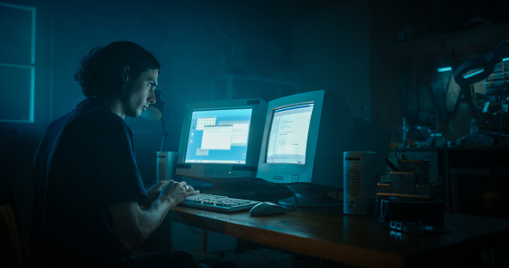 Caucasian Male Programmer Using Old Desktop Computer With Two Monitors In Retro Garage Late At Night. Evil Hacker Searching For Software Vulnerabilities, Coding DDOS Attack, Trojan Horse Program.