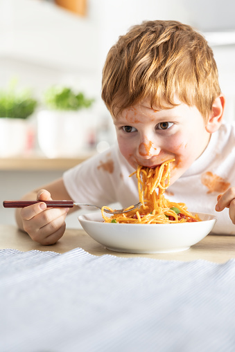 A cute little boy is eating spaghetti bolognese for lunch in the kitchen at home and is covered in ketchup.