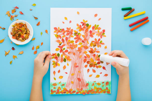 istock Little child hands gluing and creating tree shape from colorful leaves on white paper. Light blue table background. Pastel color. Making autumn decorations. Point of view shot. Closeup. Top down view. 1934876617