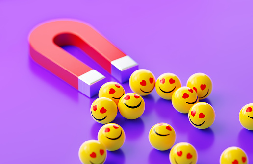 Yellow spheres textured with smiley face emoji are attracted by a red magnet on purple background. Horizontal composition.
