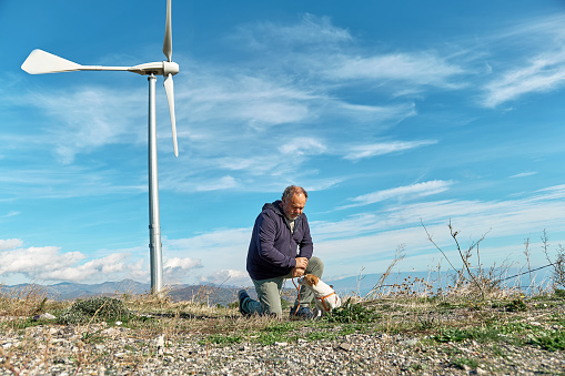 Green energy concept. Man walking with dog near mountain top industrial wind power plant, wind turbine for generating electricity in wind farm or wind park.