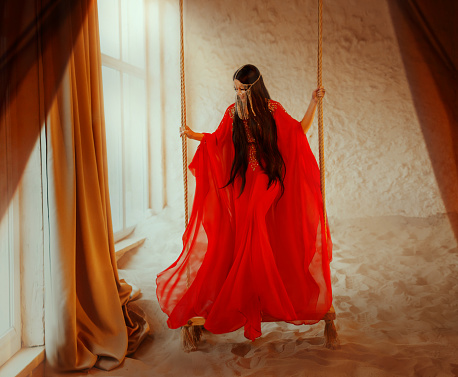 art Portrait real Fantasy girl arabic woman swinging on swing in room of full sand. red long silk abaya dress. clothes gold Mask chain Burka hide face Oriental style. desert concept of post-apocalypse