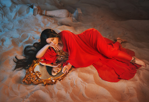 art Portrait real Fantasy girl arabic woman Fairy queen looks at reflection in mirror magic. beauty girl Lies in room full sand desert uae dubai. red long dress dream face fairy style night moon light