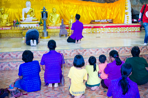 Praying thai people in cave with sleeping  buddha statue inside. Buddha's cave is in outskirts of small village Paranghmee in Phitsanulok province on temple grounds.. People are coming together for a prayer. One man is bowing in front of buddha. In foreground women, mature women and some children are sitting on their knees.
