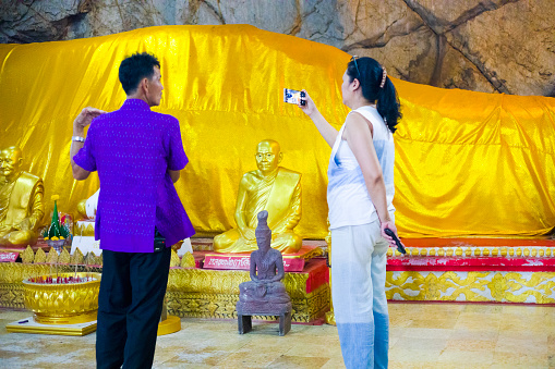 Thai man and a woman are standing  in cave with sleeping  buddha statue inside. Buddha's cave is in outskirts of small village Paranghmee in Phitsanulok province on temple grounds.. People are coming together for a prayer. Woman is holding mobile phone and is taking a video while man is talking. In front of sleeping buddha are some smaller buddha statues.