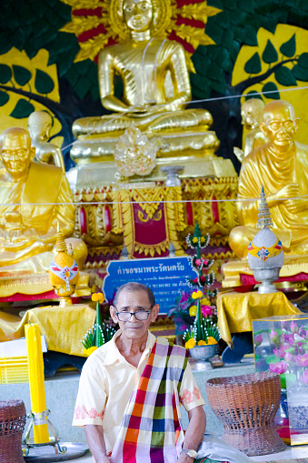Senior thai man with scarf in local temple is kneeling in front of golden buddha statues inside of temple. Temple is in small village Paranghmee  in province Phitsanulok. He has a scarf, a thai paccama around his body. Man is looking at camera. Capture taking during rural village people attend the temple for prayers. Golden buddha is surrounded by golden monk statues.