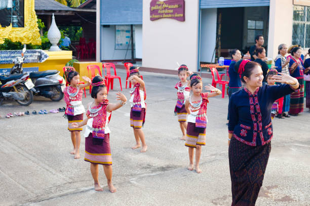 Cheerfully rum thai dancing Thai women with children in a local dance performance in a small village. Both women and children are dressed traditionally. The children  are dancing barefoot. Scene is in small village Paranghmee in province Phitsanulok. In background is a local building. In right background are some people attending performance. At left background are motorcycles and part of image of king