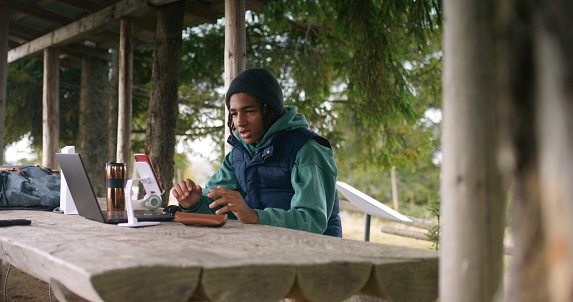African American young boy sits in wooden gazebo, talks with teacher at video call using laptop and phone on tripod. Tourist remotely studies during holidays in mountain forest. Outdoor study concept.