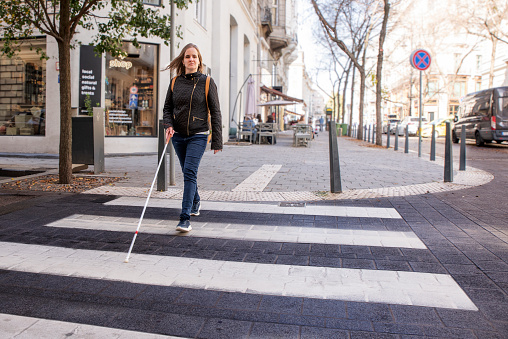 Portrait of blind woman with white cane crossing on the road in the city. A visually impaired woman wearing casual clothes and using her cane to cross the street. Full length shot.