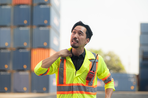 Portrait of an Asian male worker wearing a safety vest and hard hat, taking a break from work with a container box in the background