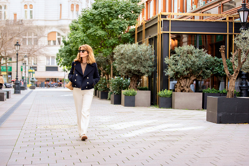 A middle-aged woman with blonde hair is walking down the street in the city centre. Attractiv female wearing sunglasses and blue coat. Full length shot.
