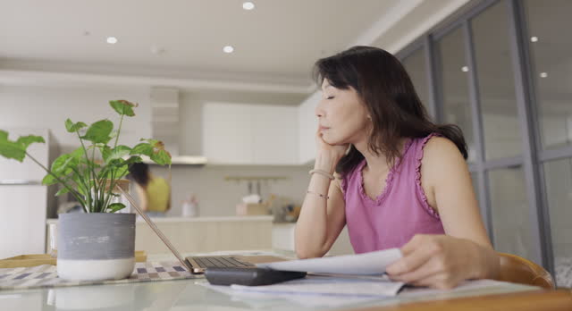 A woman is at home, holding a collection of paper bills and financial plans. She appears to be focused on managing her personal finances, possibly sorting through bills, budgeting, or organizing her financial matters