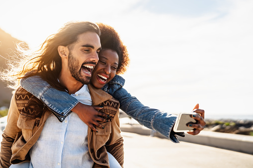 Happy multiracial couple having fun taking selfie with mobile smartphone during vacations - Youth people love and social relationship concept