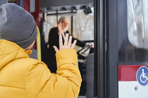 Back view of little boy in orange jacket and grey cap saying goodbye to blur driver after travelling in public vehicle. Crop of child male waving to driver while getting off bus. Concept of city life.