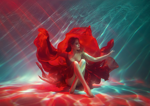 Calm relaxation meditation concept. Sexy fashion model Fantasy woman sitting under water sea, red long silk dress fabric floating. fairy girl posing in deep pool underwater shooting Art Magic light