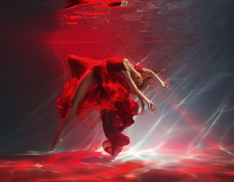 fashion model posing with sexy legs. Fantasy woman dancing under water sea red long silk dress fabric floating. Belerina fairy girl dancer swims in dark deep pool underwater shooting Art Magic light