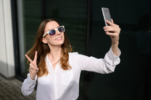 A woman in a white shirt is talking to a friend on the camera phone while traveling in a new city and shows a sign on her fingers that everything is fine.Travel, blog and blogger concept.